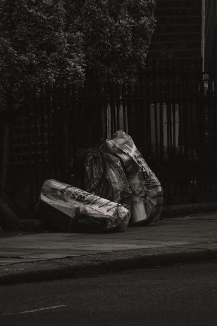 Three large black plastic rubbish bags filled with waste, leaning against a dark metal fence on a pavement in a residential area, with a lush leafy tree partially visible above the fence and the faint