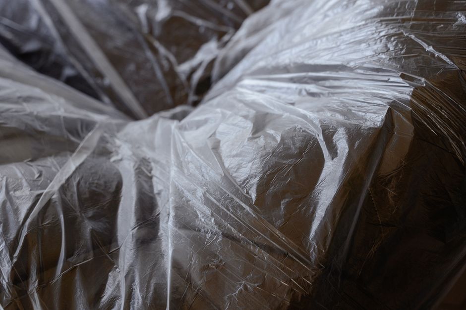 Three large black plastic rubbish bags filled with waste, leaning against a dark metal fence on a pavement in a residential area, with a lush leafy tree partially visible above the fence and the faint