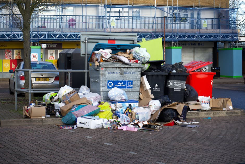A collection of overflowing waste bins and scattered rubbish situated on a paved area in front of a retail or commercial space, with a grey metal bin labeled for mixed paper and cardboard in the foreg