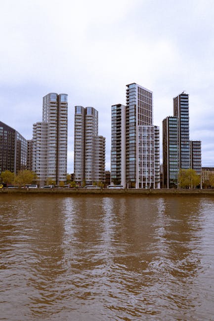 A cityscape featuring a cluster of modern high-rise buildings situated along a riverbank, with reflections visible on the water's surface. The buildings vary in height and architectural style, with so