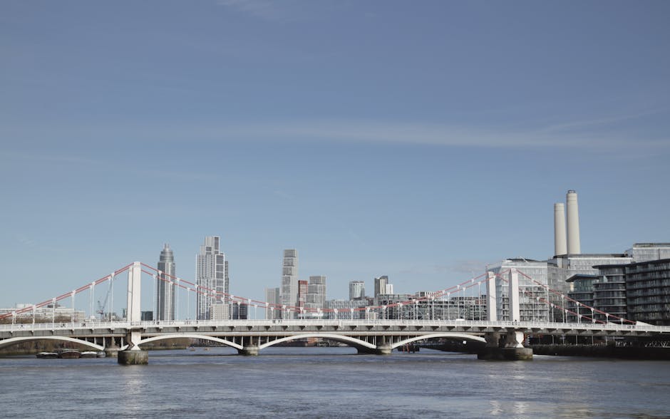 A cityscape featuring a cluster of modern high-rise buildings situated along a riverbank, with reflections visible on the water's surface. The buildings vary in height and architectural style, with so